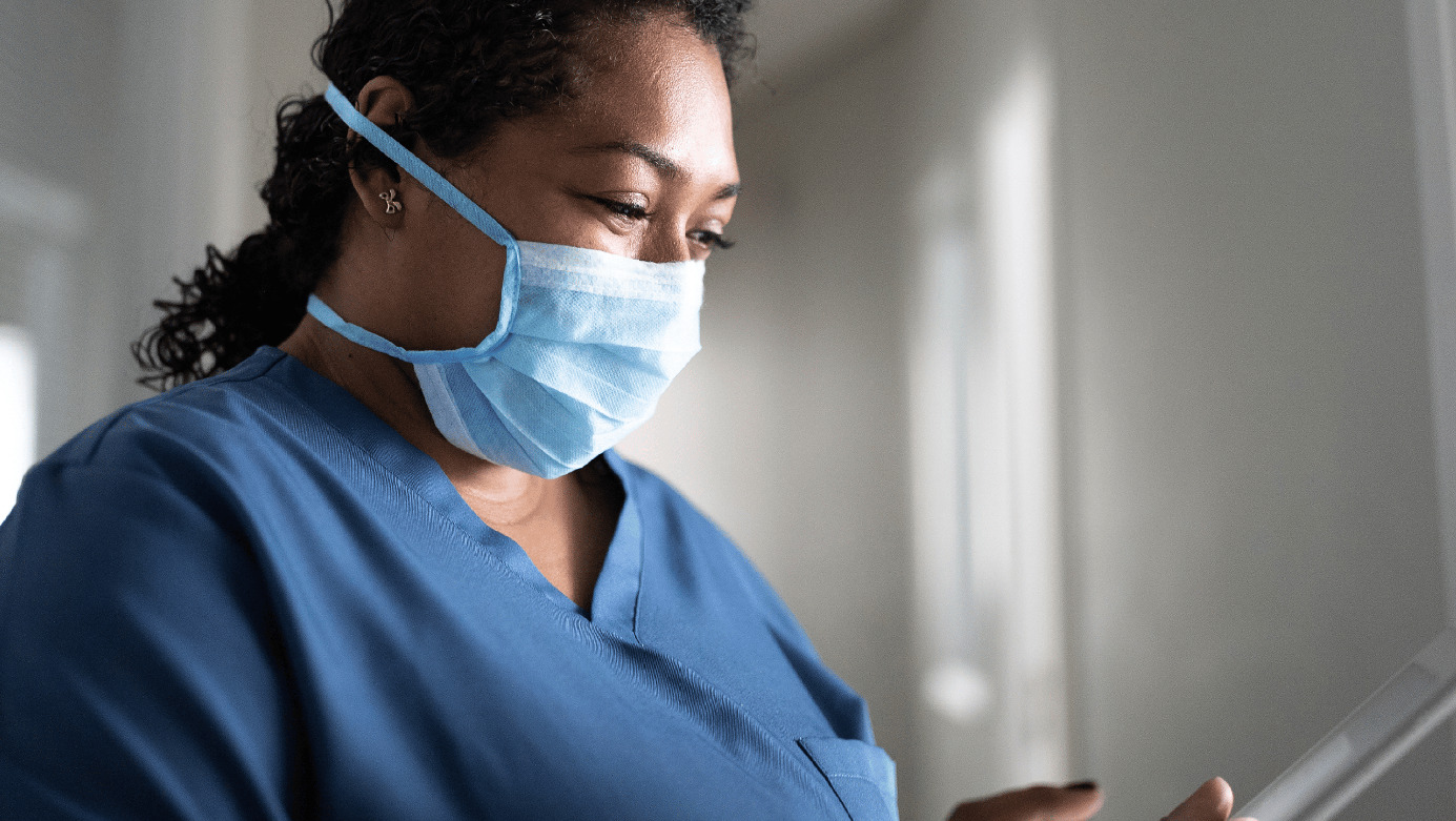 Masked nurse in blue scrubs checking patient records on tablet